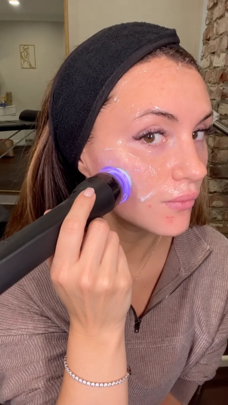 A woman in a headband gliding the pen over gel on her cheek with the blue light on
